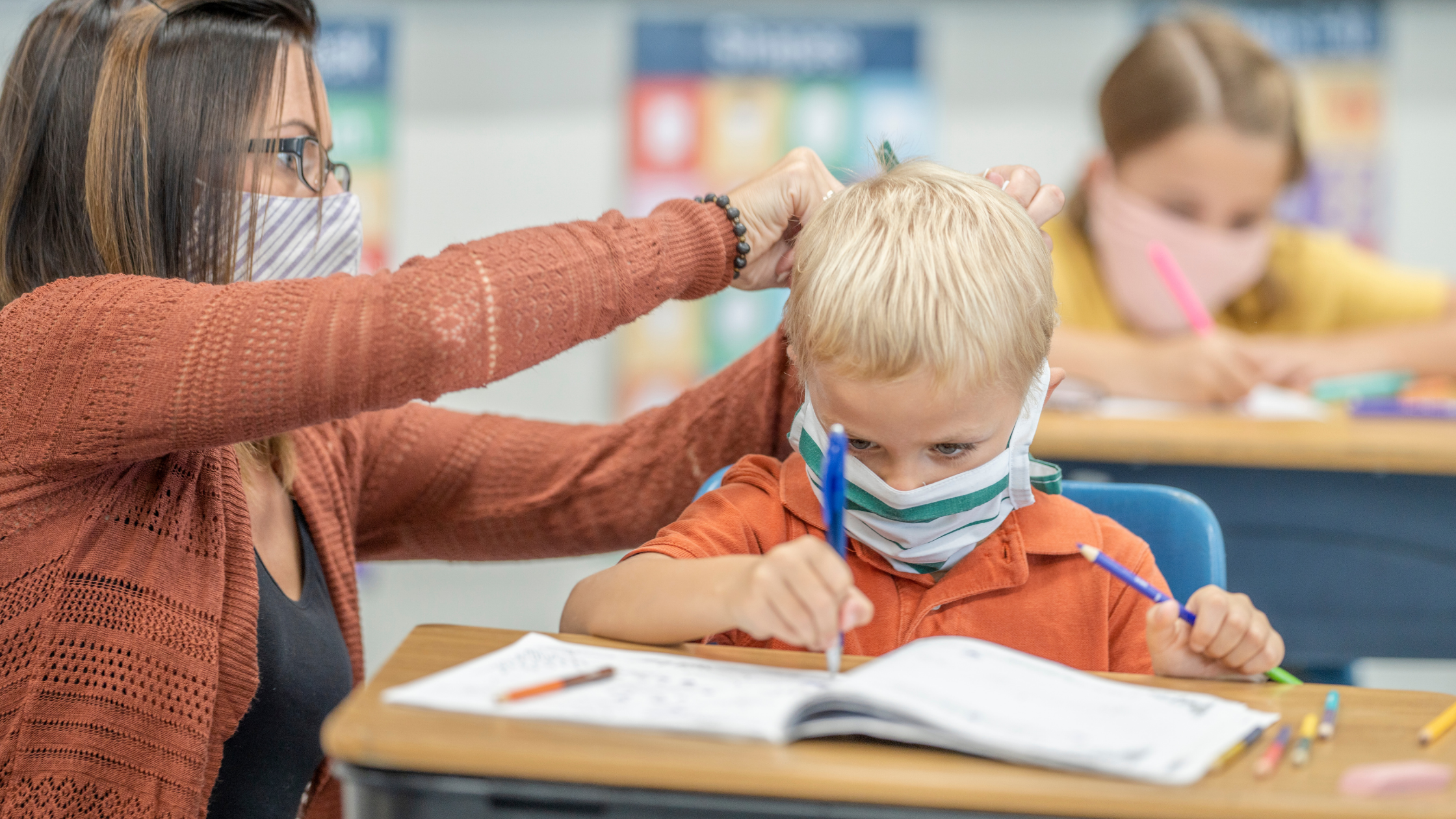 a white fe3male teacher with medium length brown hair crouches next to the desk of a blonde boy as he writes in an open workbook. Both the teacher and student wear masks. She reaches behind his head, presumably fixing his mask.
