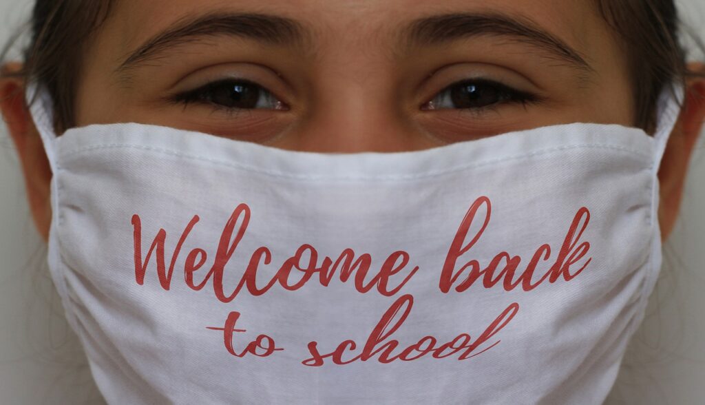 A young girl smiles beneath a mask, her dark brown eyes and wisps of her dark hair the only parts of her face we see. Her white mask has "Welcome back to school" written on it in red loopy letters.