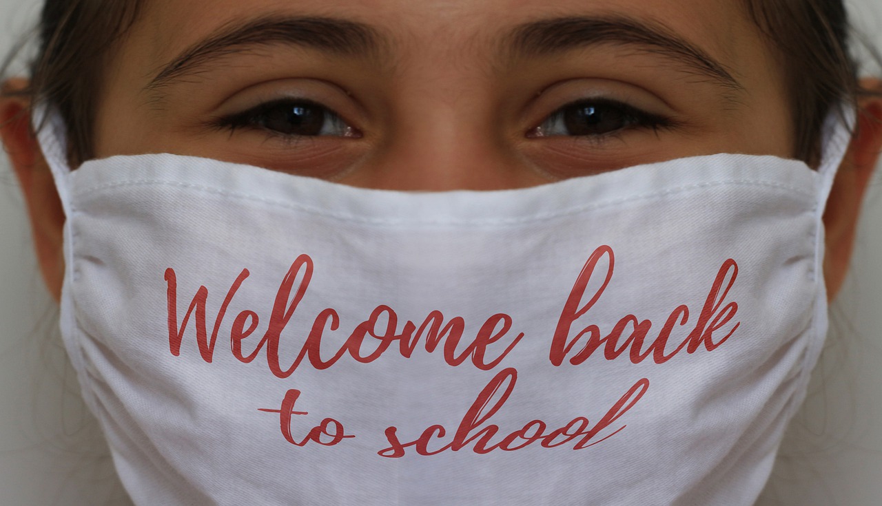 A young girl smiles beneath a mask, her dark brown eyes and wisps of her dark hair the only parts of her face we see. Her white mask has "Welcome back to school" written on it in red loopy letters.