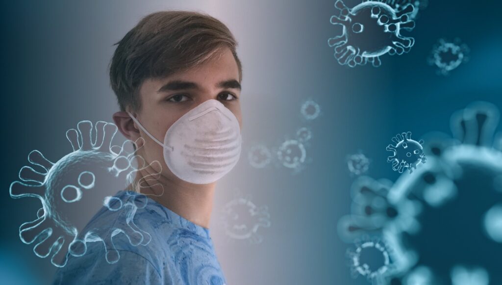 A white male teenager stands to the left of the photo, his body at a sideward angle with his head turned toward the camera. He wears a white mask. A blue overlay with coronavirus graphic covers the image.