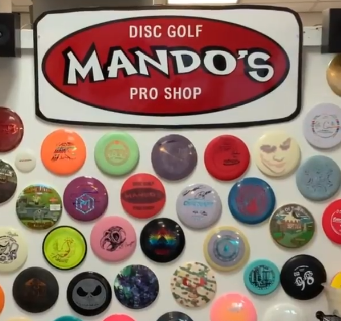 A wall covered with colorful round discs below a sign that reads "Mando's" with Disc Golf above it and Pro Shop below it. The white text is within a dark red oval.
