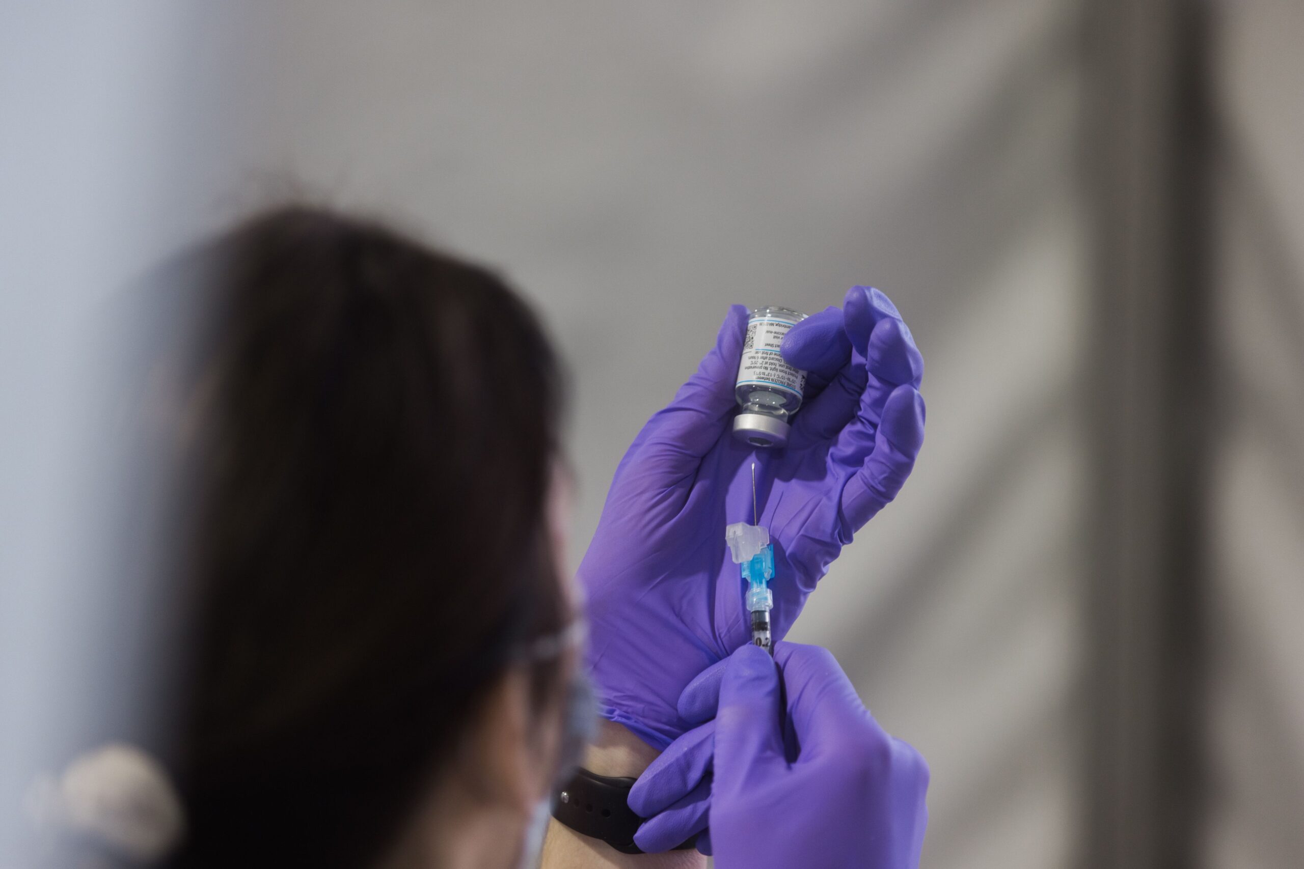 An individual wearing a mask prepares a new COVID-19 vaccine. The individual, appearing to be a brunette female, has their back to the camera