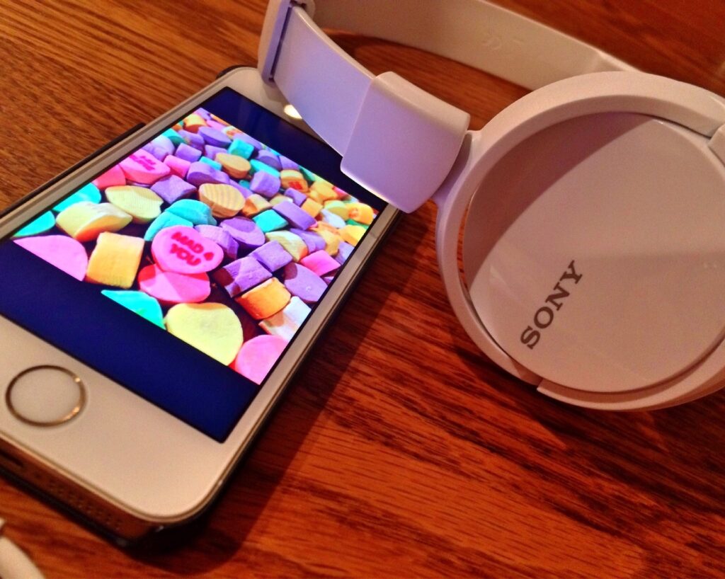 a white cell phone and sony pair of large round headphones sit on a wood tabletop. On the screen of the iPhone are colorful candy hearts.