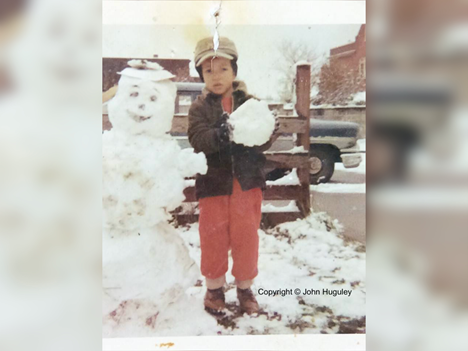 In an old photograph, a Black child stands next to a snowman that matches his size. He holds a large snowball. He wears a red jumpsuit and dark coat. A oversized winter hat tops his head.