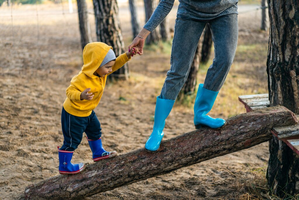 An adult and child, both wearing rubber rainboots, balance on a slanted log in the woods. The adult holds the hand of the child.