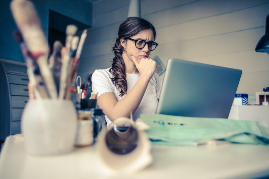 A brunette female wearing thick black rimmed glasses sits at a desk in front of a laptop. The Photo is taken from behind and to right of the laptop, capturing the female who looks thoughtfully at the screen with her fist to her chin. Blurred in the foreground are papers, a cup of pens and paint brushes, and other office supplies on the desk.