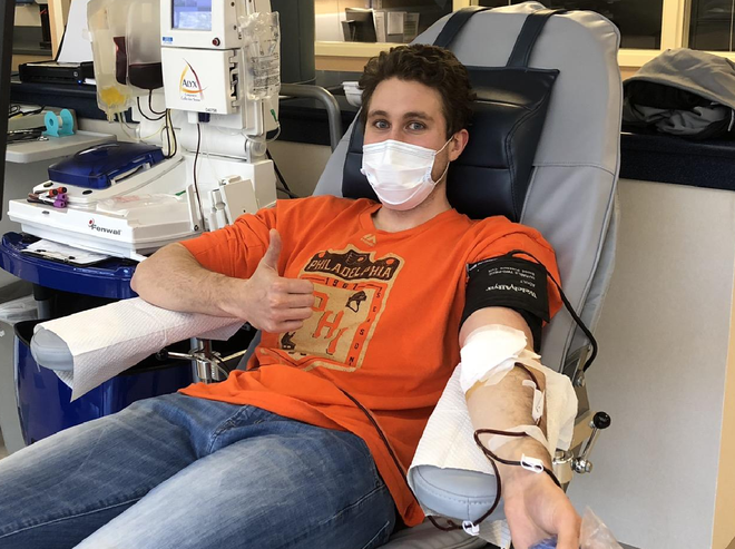 a teenage male sits in a chair with his arm extended, medical tubing extending from his arm to a machine behind him. He is donating platelets.
