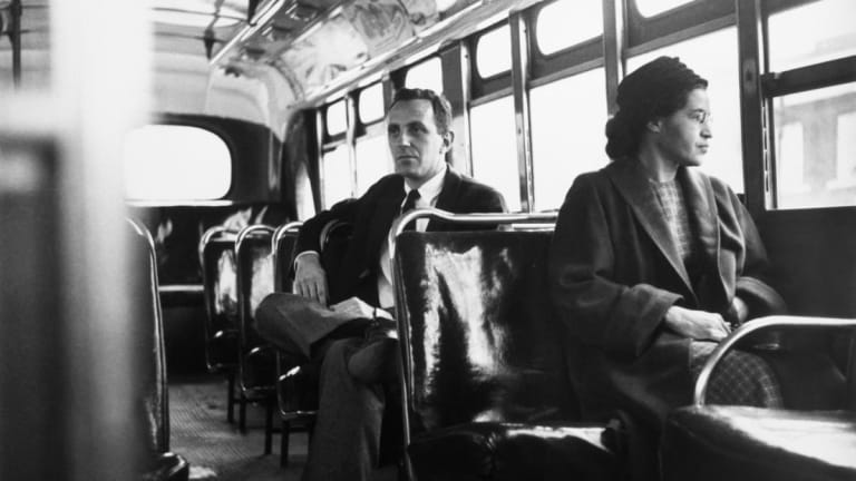 A black and white photo of Rosa Parks, an African American woman wearing a long coat and round hat, sits on a public transit bus looking out the window. The bus is empty beyond a single white man in a suit that sits behind her.