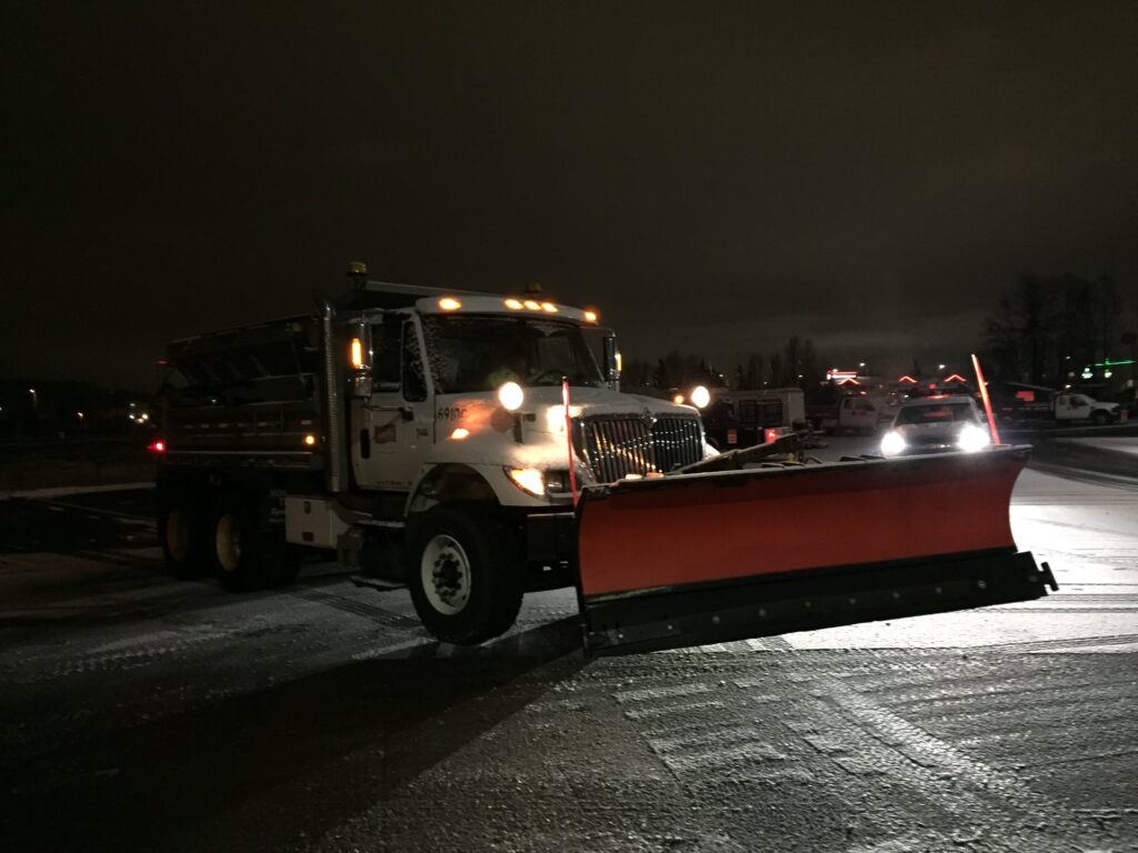 A City of Auburn snow plow on a snowy winter night. The plow is parked at an angle with its lights on, a second plow illuminates it from behind.