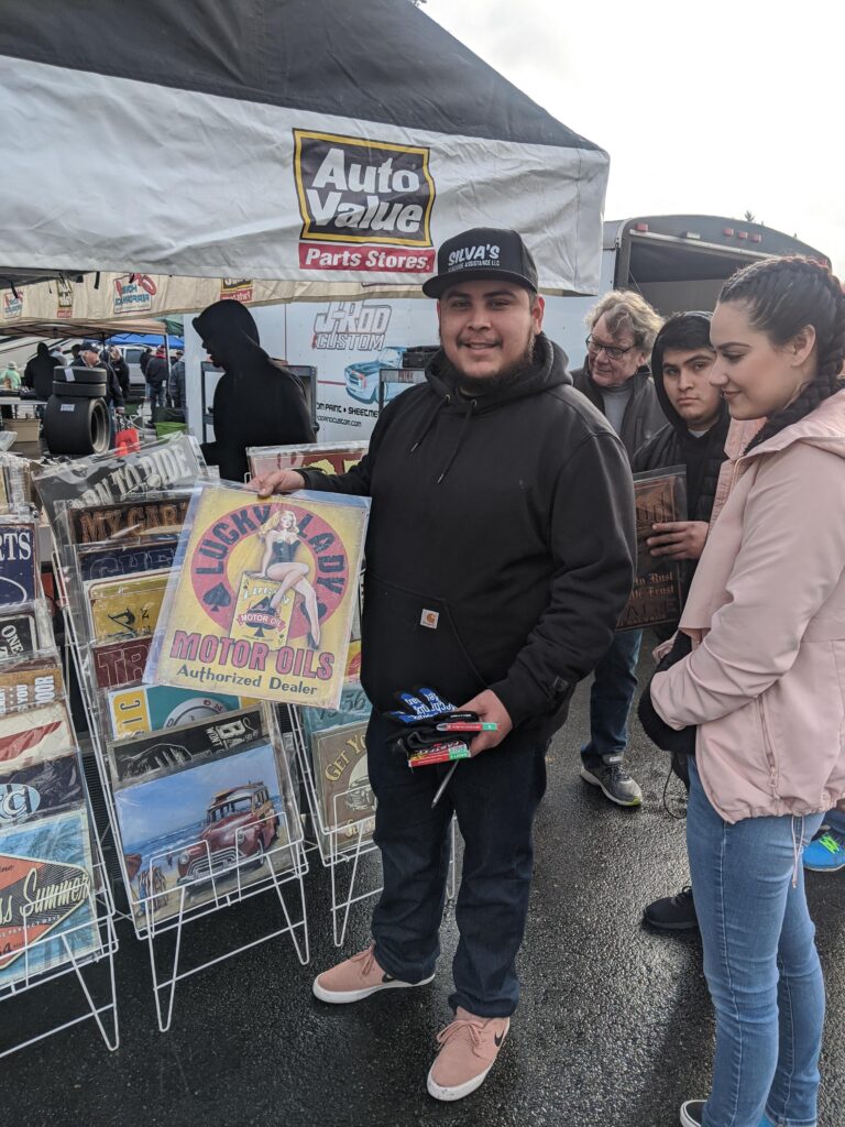 A man, smiling, holds up a lucky Lady motorsports sign in front of a vendor tent