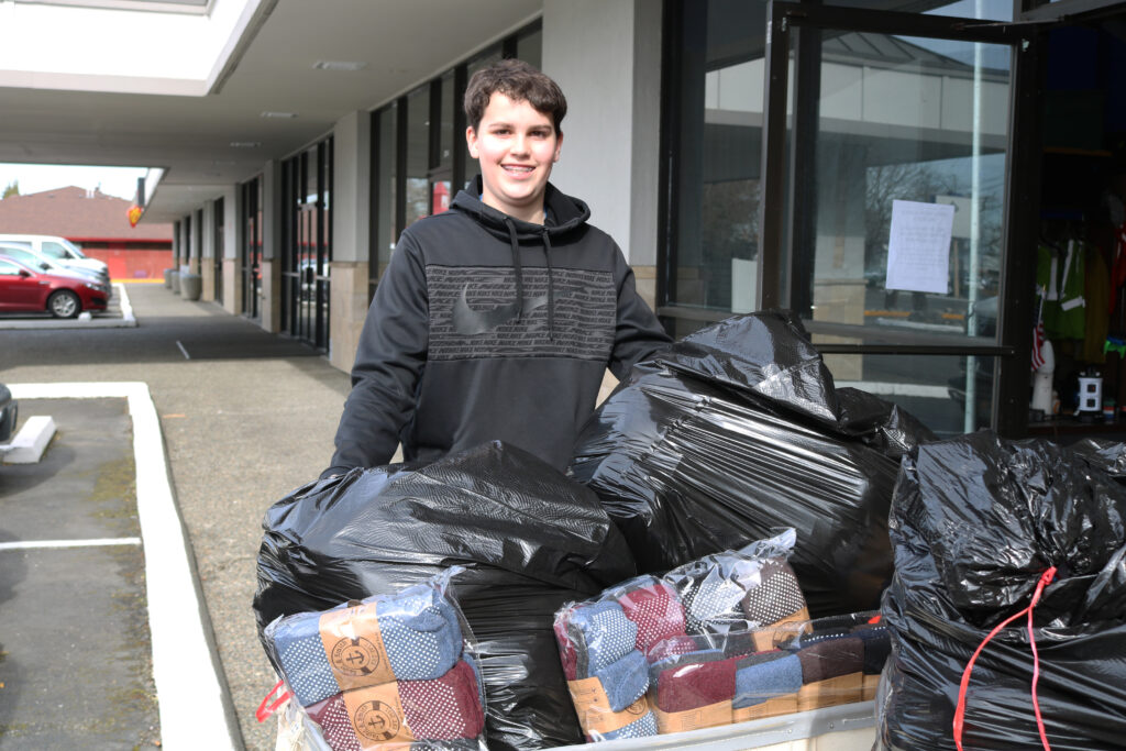 Boy Scout Alec Mayer stands in front of a wheeled laundry tote overflowing with black garbage sacks full of socks.