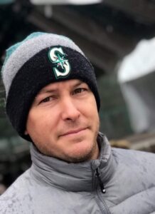 A white male in a gray winter jacket wearing a gray, teal and dark blue Seattle Mariners beanie