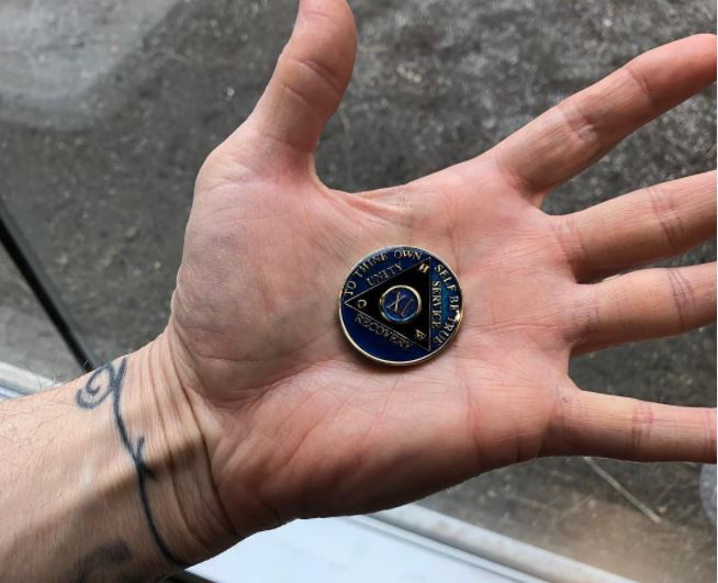 A person holds an 11 year AA sobriety token in the palm of their sprawled open hand. Their wrist has a thin black tattoo. olds an eleven year AA sobriety token in its palm. On the individual