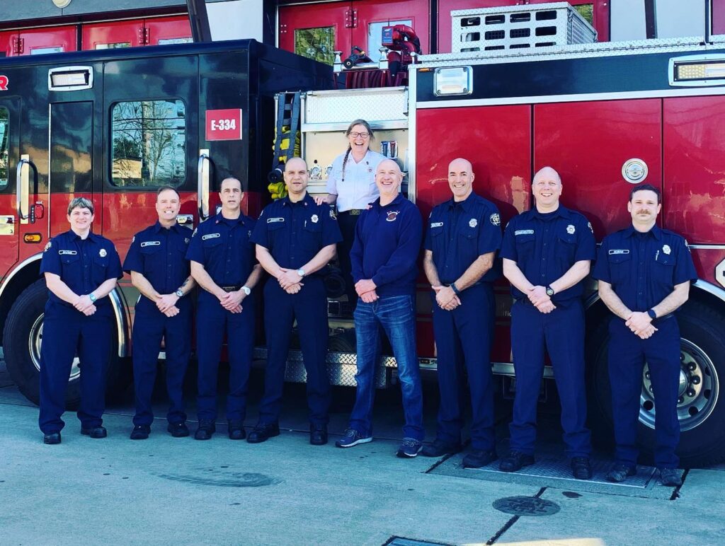 8 individuals in dark blue VRFA uniforms stand in front of a fire truck. Between them stands a female in a white shirt. 