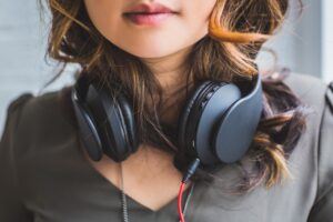 A pair of large round headphones wrap around a female's neck, her auburn hair scrunched under them. The photo is from her lips down to her chest.