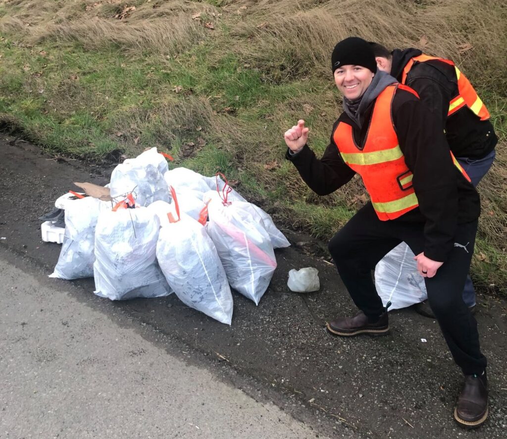 Two adult males in orange safety vests stand next to a pile of full trash bags. The male in front is blocking the second male as he leans toward the pile of bags holding his hand up making a "just a little" gesture with his hand.