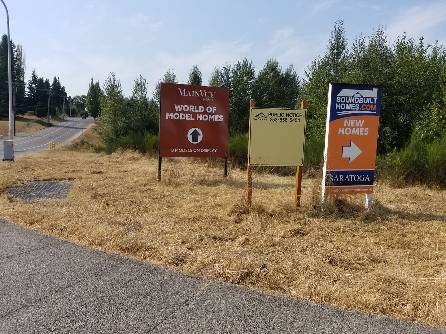 Three developer signs on an undeveloped corner. The middle sign states "Public Notice" from the City of Kent, but holds no notices.