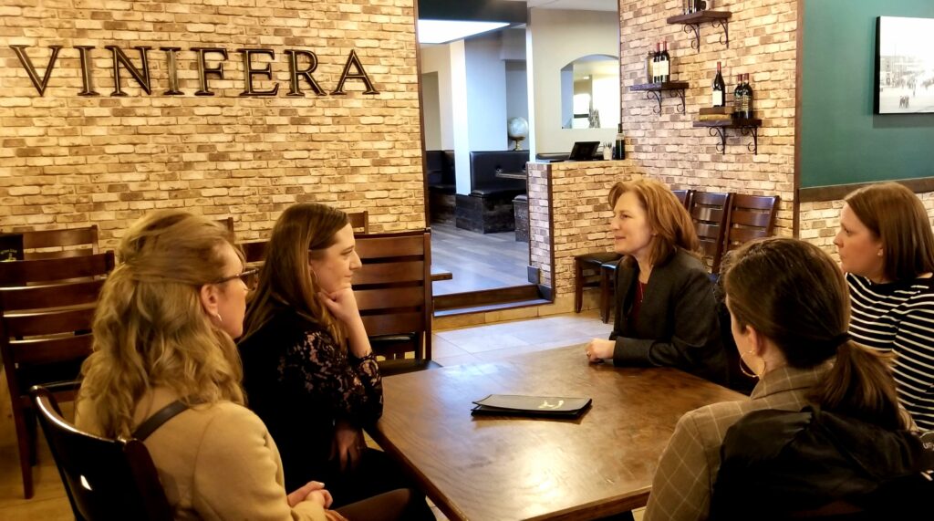 Rep. Kim Schrier and Brittiany Karlson sit across from each other at a table in Vinifera. They appear mid-conversation, with three other women listening
