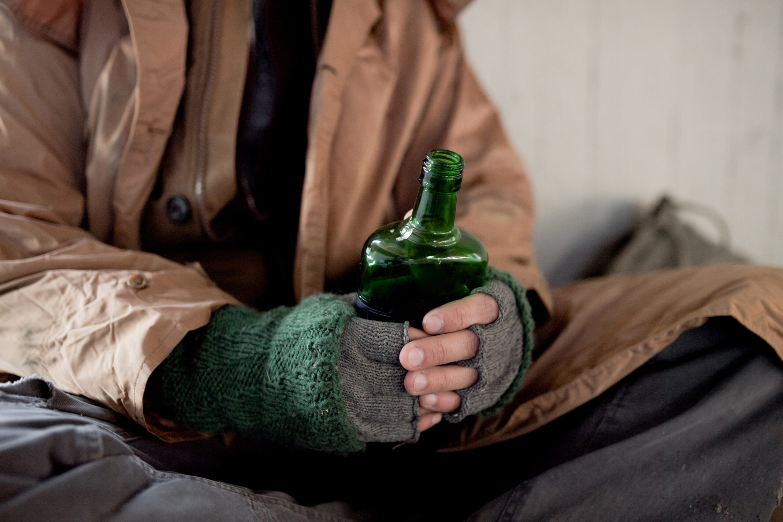 A midsection view of homeless man sitting outdoors in city asking for money donation, holding a bottle of alcohol.