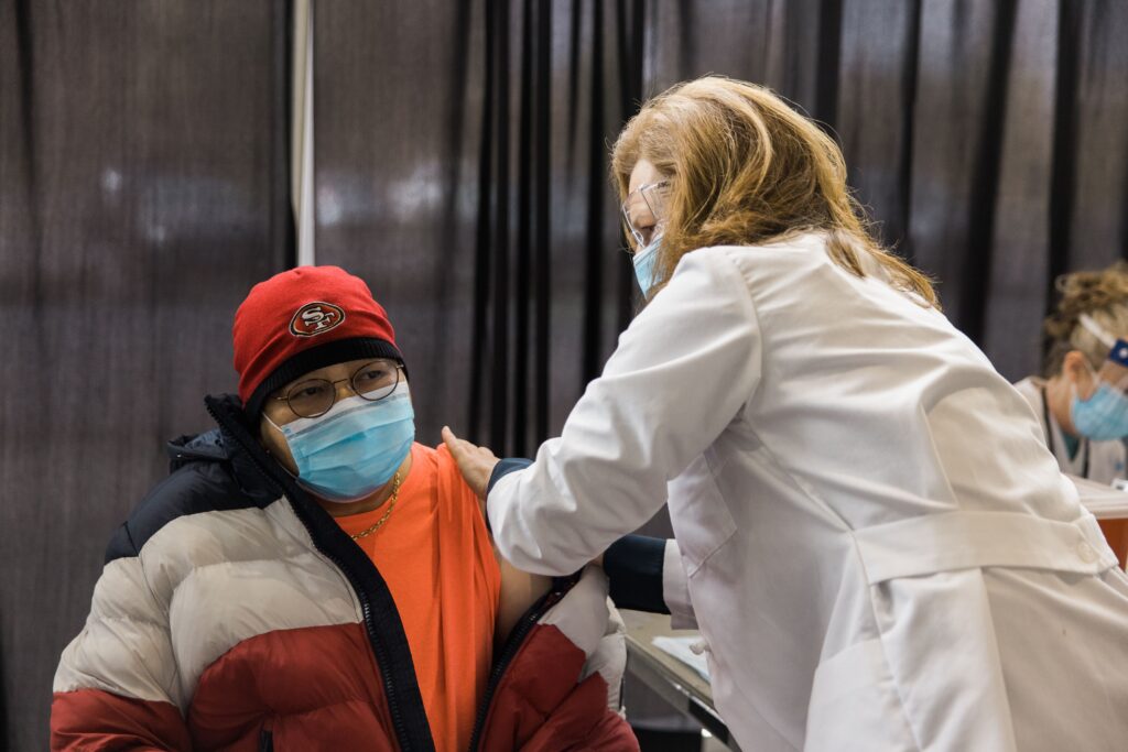 A female doctor in a mask, eye protection and a white lab coat administers an injection to a male in a neon orange t-shirt, gray and red puffy winter coat and SF 49ers beanie. The male wears a mask and glasses, his coat unzipped and partially pulled down his arm to allow for the injection.