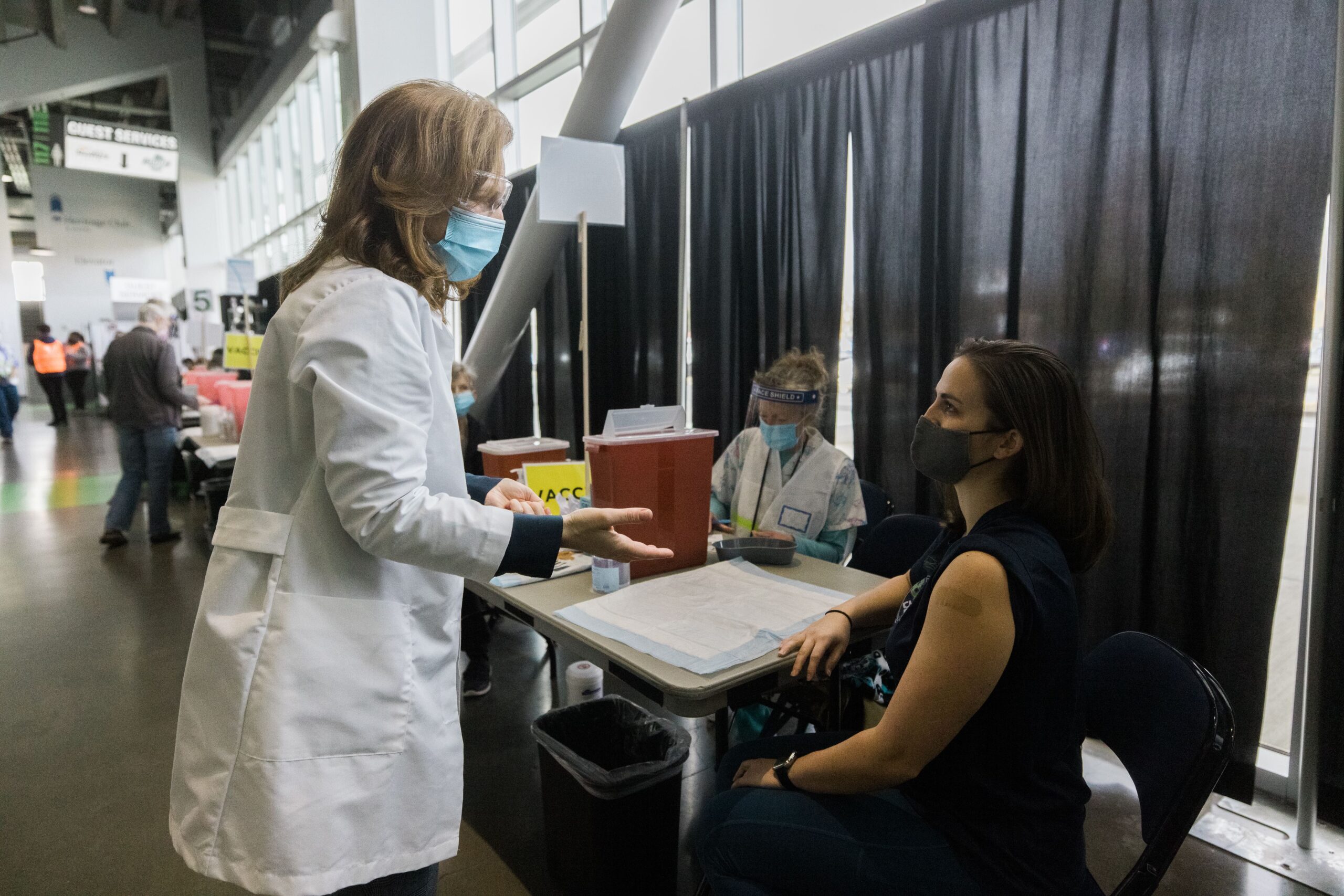 A female doctor in a white lab coat speaks to a female sitting in a chair. They are in