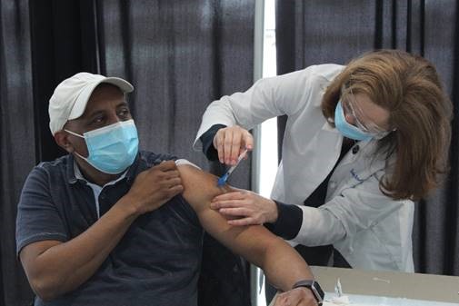 A female doctor in a mask, eye protection and a white lab coat administers an injection to a male in a blue t-shirt, white baseball hat and mask. The man holds his sleeve up to allow for the injection.