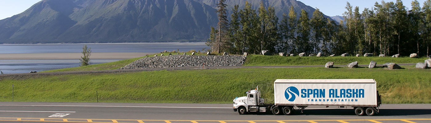A Span Alaska tractor trailer drives on a high way with a serene mountainscape and lake background