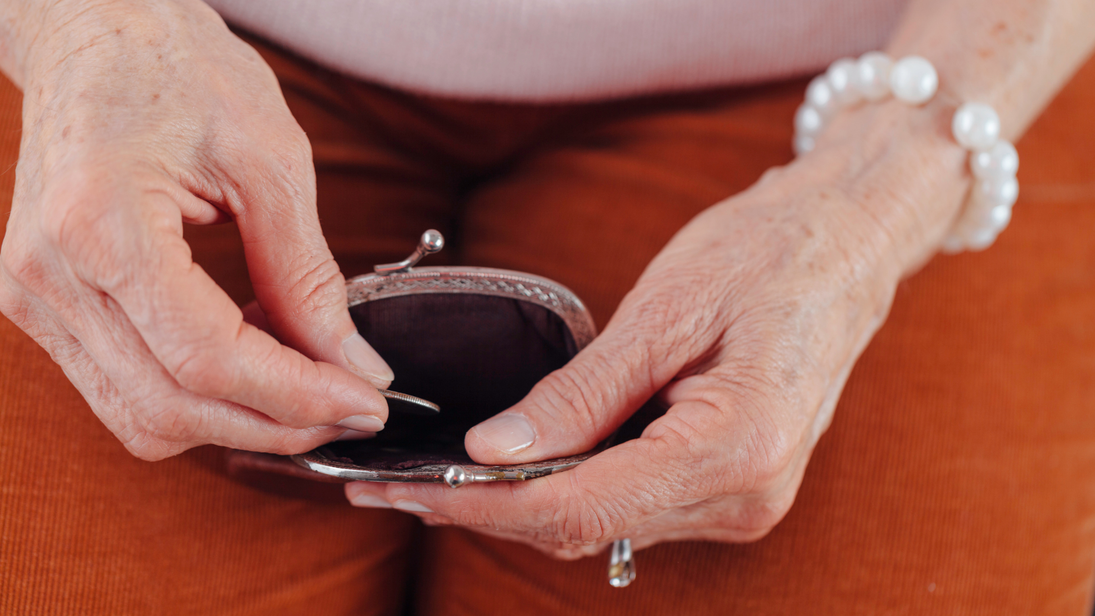an older woman holds open a change purse on her lap. We see only her hands, with one putting a coin into the purse