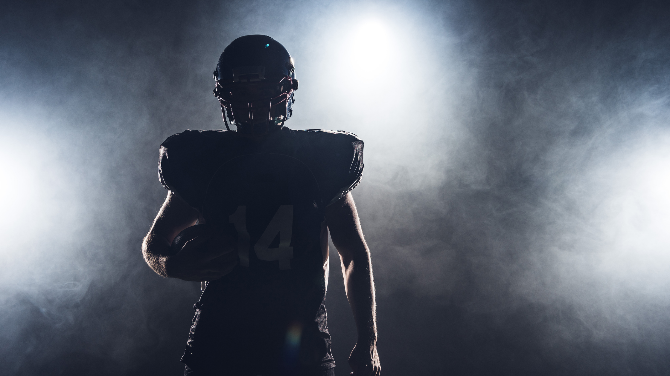 The silhouette of a football plater is surrounded in a thing smoke, shielding his features. He cradles a ball in his left arm.