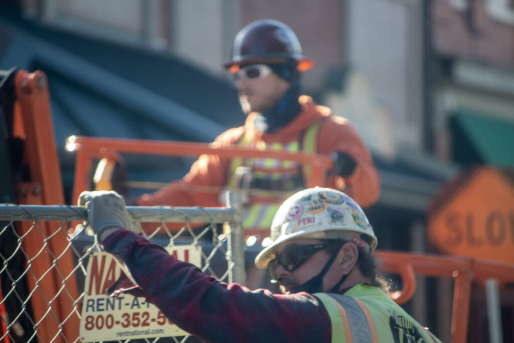 A construction worker in a hard hat and safety gear stands with his hand on top of a tall chainlink fence that surrounds a construction site. A second worker is seen in the background working.