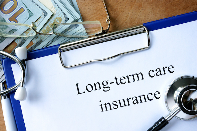 A clipboard with "Long-term care insurance" written on a paper clipped on it, a stethoscope resting toward the bottom. A stack of 100 dollar bills sits in the upper left-hand corner.