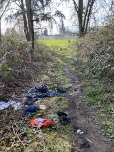 A worn path into the woods with discarded clothing and trash along it, beyond the woods appear gray buildings.