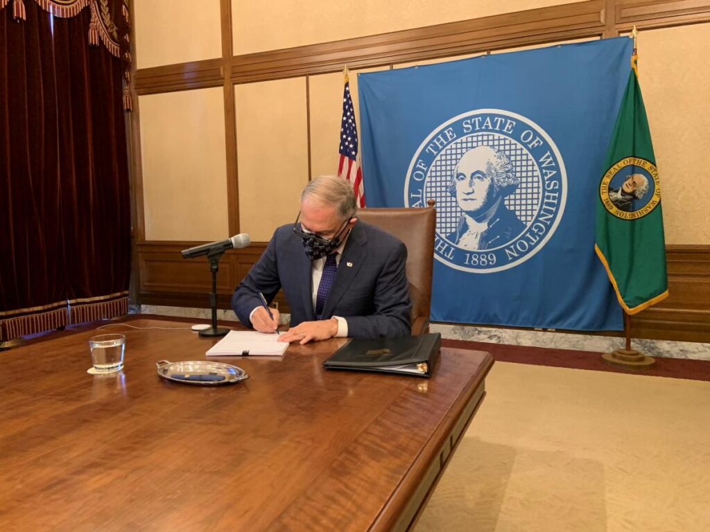 WA Governor Jay Inslee, wearing a mask and sitting alone at the end of a long table, signs a bill into law