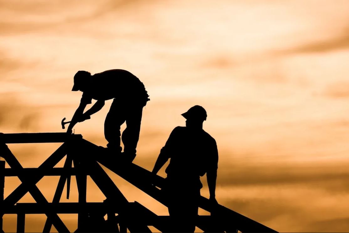 twoconstruction workers work on top of the frame of a home. The workers are silhouettes to a vibrant orange sky.