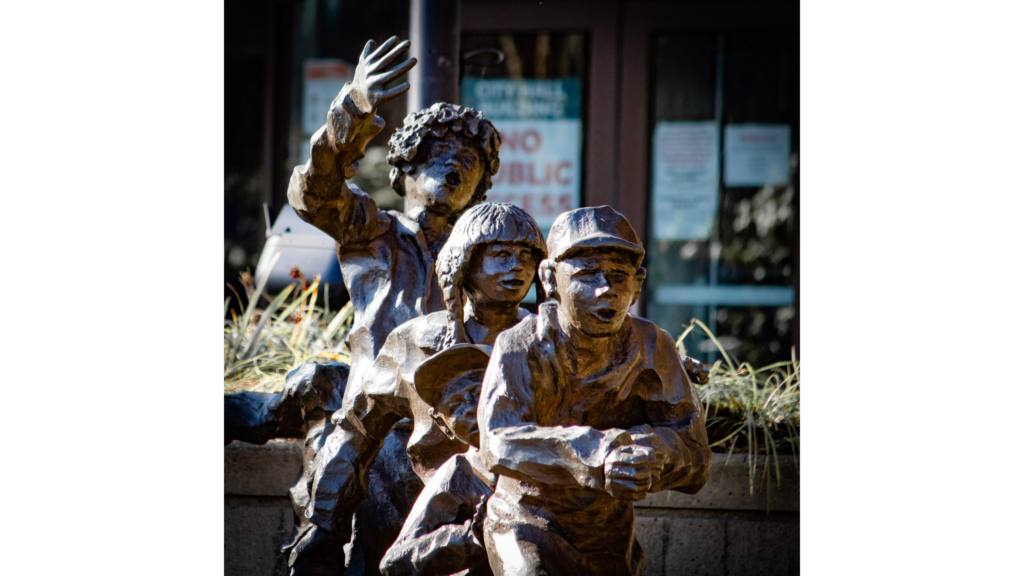 A metal statue of 4 children in a line playing. The statue is in front of Auburn City Hall, which can be seen out of focus in the background.