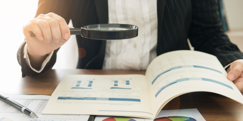 an individual holds a magnifying glass over a financial report booklet, additional pages of reports on the desk below the booklet