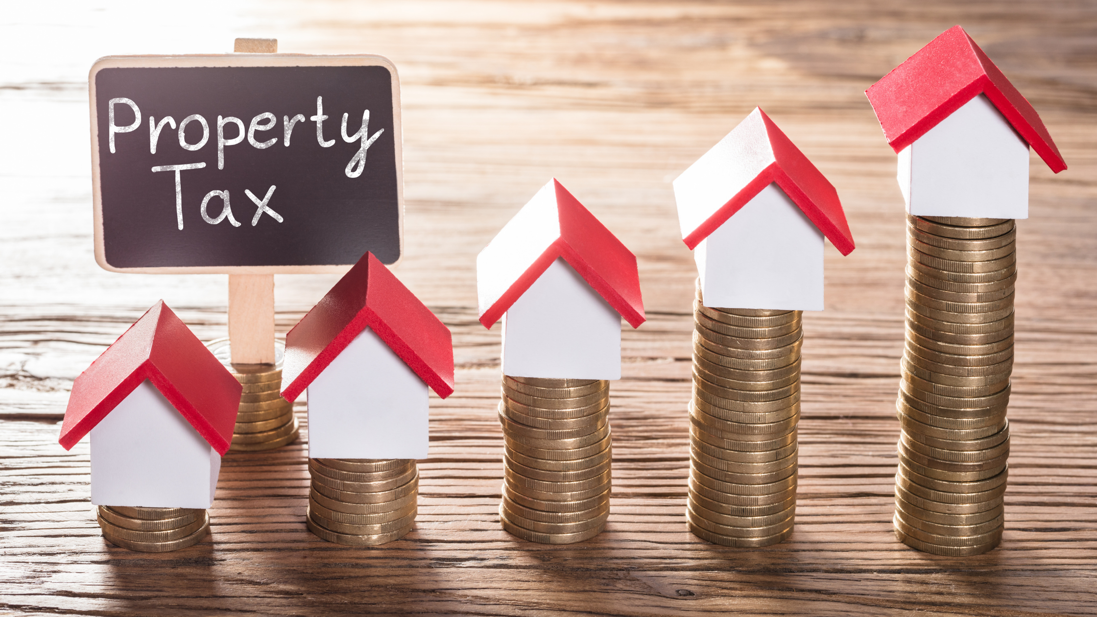 small houses sit on stacks of coins that increase in size. A "property tax" sign sits near the lowest stacks.