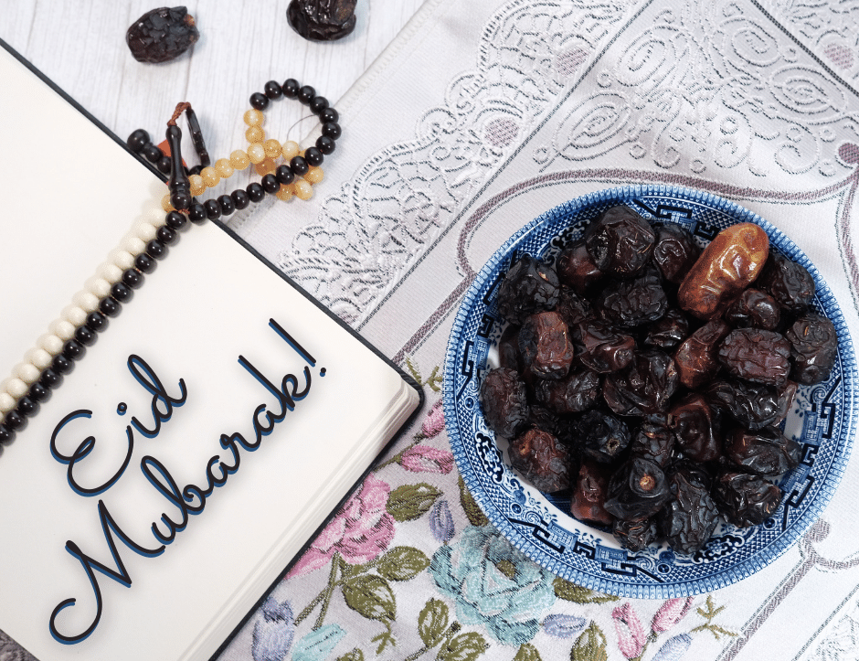 An open prayer book sits on a prayer rug with prayer beads and a bowl of dates next to it. Eid Mukarak! is written on the page of the book.
