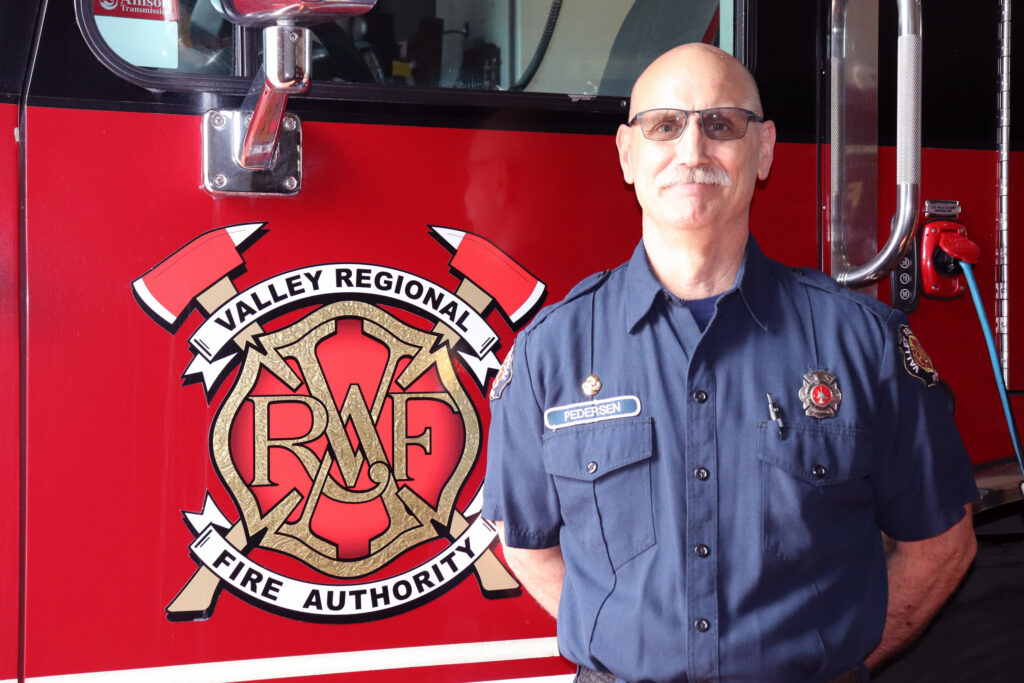 VRFA Firefighter Neil Pedersen stands with his hands behind his back in front of a VRFA fire engine.