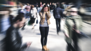 A woman stands with her hands over her face among a crowd of people on a busy sidewalk. The crowd is blurred, the woman the only thing in focus