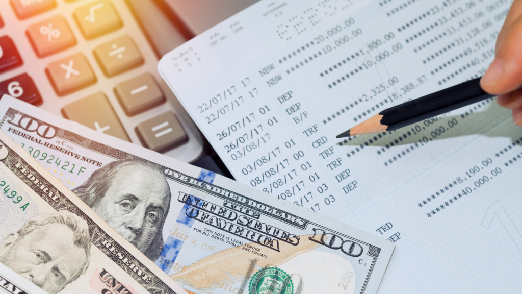 two hundred dololar bills lay over a bank accounting sheet, a person hovering a pencil as the reconcile the sheet