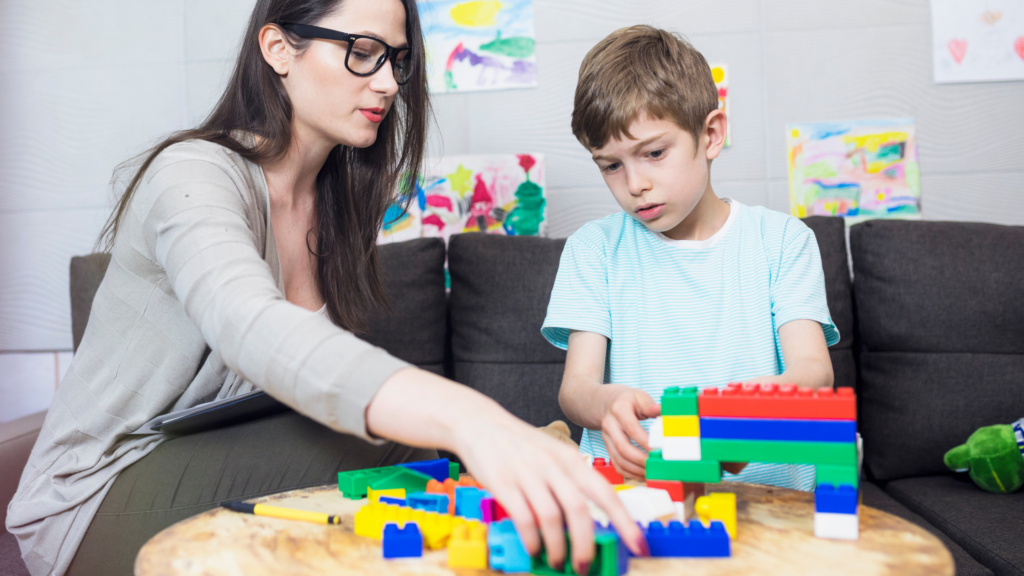an adult female sits at a table with a young boy, playing with lego blocks in a professional or educational setting. A clip board sits in the woman's lap implying this is an evaluation or therapy session.