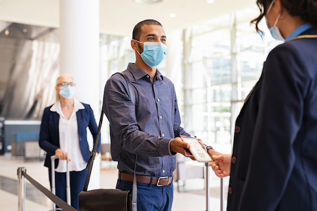 Indian passenger wearing surgical mask showing e-ticket to flight attendant at boarding gate. Young mixed race businessman showing boarding pass on mobile phone to air hostess while wearing protective face mask during covid pandemic. Multiethnic business man in a row with flight reservation hand the phoone to stewardess at airport keeping social distance.