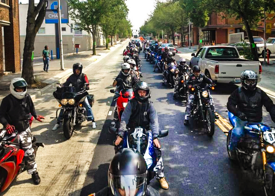 two long rows of motorcycles line a city street, ready to ride. Two motorcycle guides are to the left of the group.