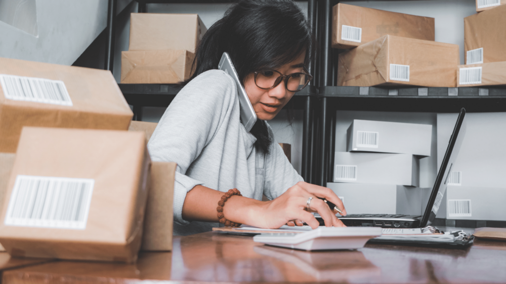 a woman sits at a desk on the phone, a laptop in front of her packages on the desk and on a shelf behind her.