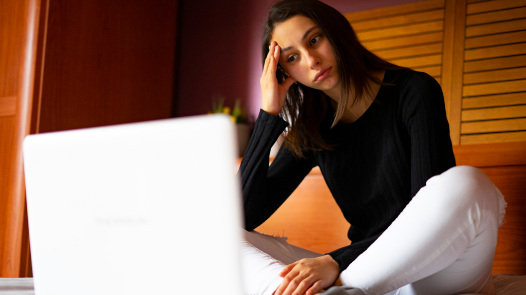 A young woman sits cross legged in front of a laptop looking dejected, her hand on her forhead