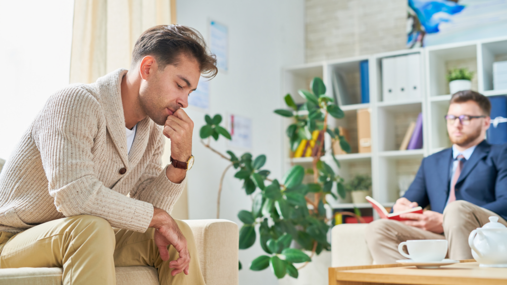 A man sits in a chair looking pensive, out of focus in the background another man listens with a notebook open on his lap. They are in a brightly lit office. It is implied this is a therapy session.