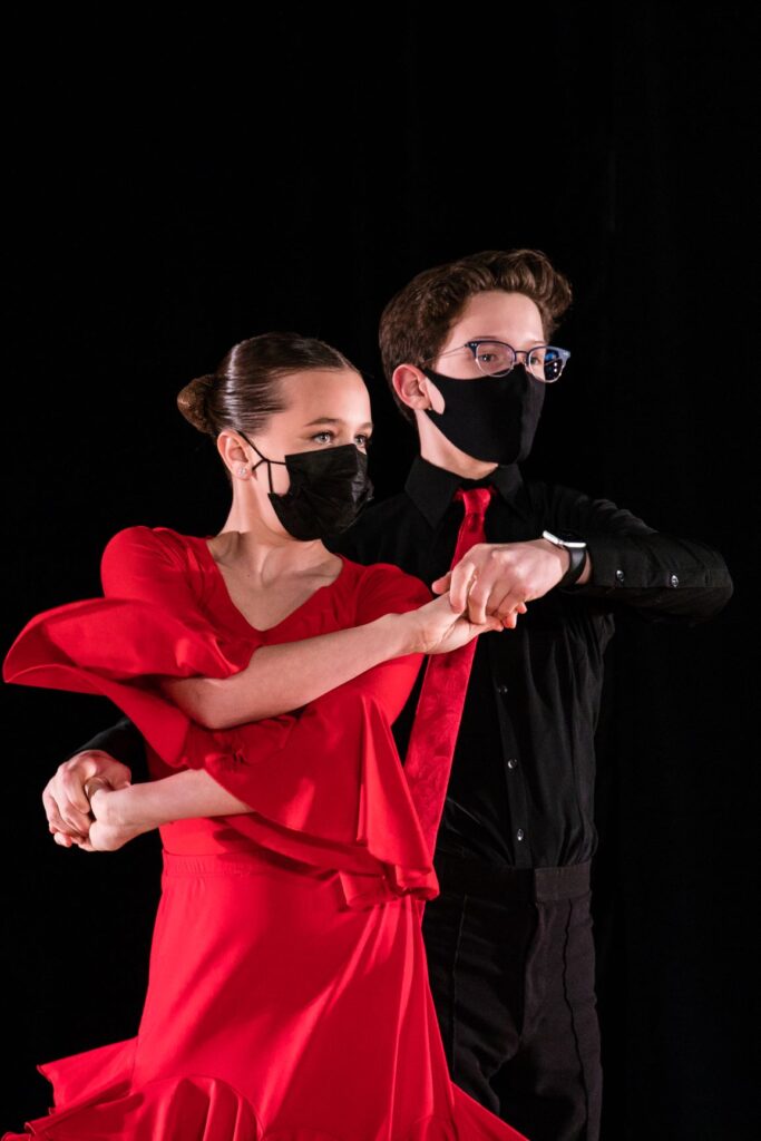 A couple are captured mid-dance. The female wears a red dress, the male a black shirt and pants and red tie.