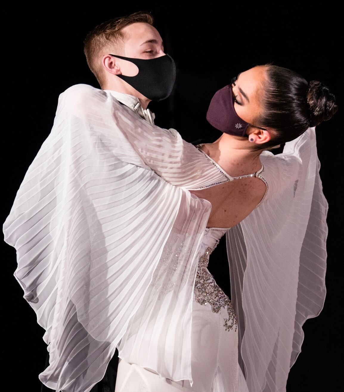 Two ballroom dancers embrace with boxed posture. The female of the pair wears a white dress with oversized bell sleeves that drape over the pair.