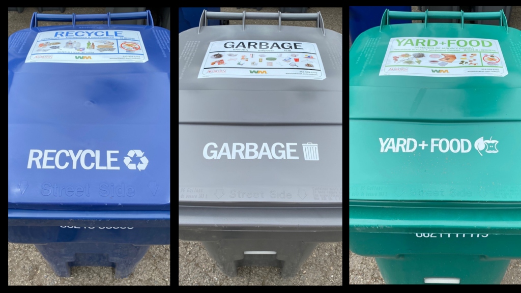 The tops of three Waste Management containers in a row. A blue container labeled Recycling, a Gray container labeled Garbage, and a green container labeled Waste.